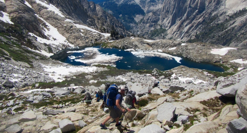 A group of people wearing backpacks hike near an alpine lake, framed by mountains. 